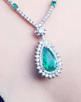 A close-up of a teardrop-shaped emerald pendant necklace with a double halo diamond, worn on a person's neck. The necklace chain also features small emeralds.