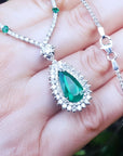 Pear cut emerald necklace with double diamond halo on 18K white gold chain, displayed on a person's hand.