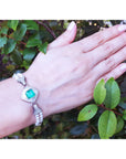 Hand wearing a pearl bracelet with a square emerald against a leafy background.