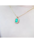 Emerald pendant with a halo of diamonds on a gold chain, worn on a person's chest.