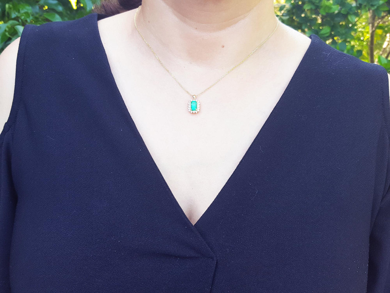 A woman wearing a blue top with a necklace featuring a natural emerald against a blurred green background.