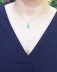 A woman wearing a blue top with a necklace featuring a natural emerald against a blurred green background.