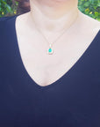 A woman wearing a dark blue top with a necklace featuring a natural emerald pendant against a blurred green leafy background