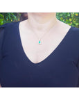 A woman wearing a dark blue top with a necklace featuring a natural emerald pendant against a blurred green leafy background