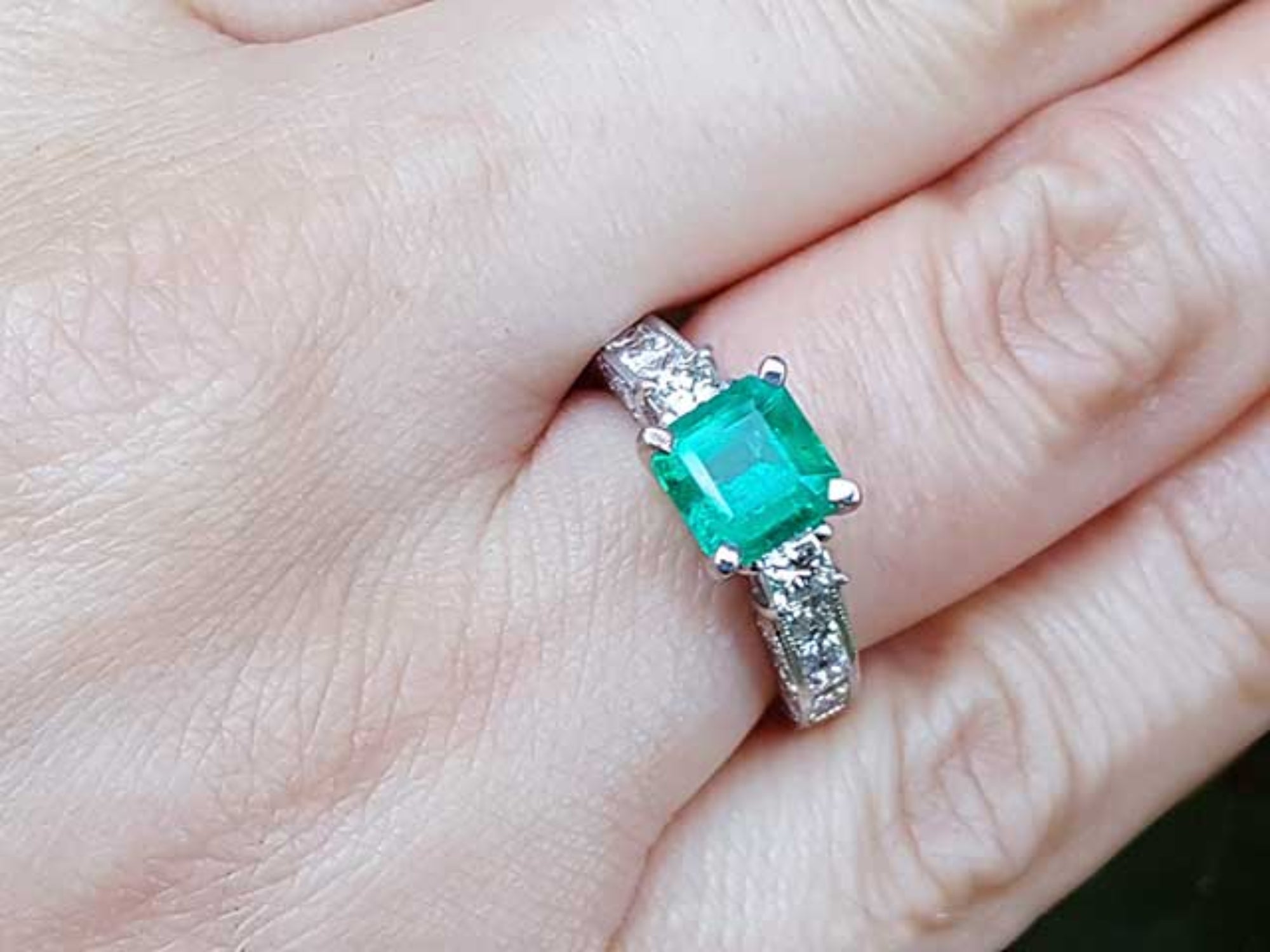 A square emerald engagement ring with princess cut side diamonds on a person's hand.