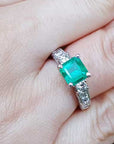A square emerald engagement ring with princess cut side diamonds on a person's hand.