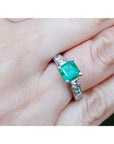 A square emerald engagement ring with princess cut side diamonds on a person's hand.