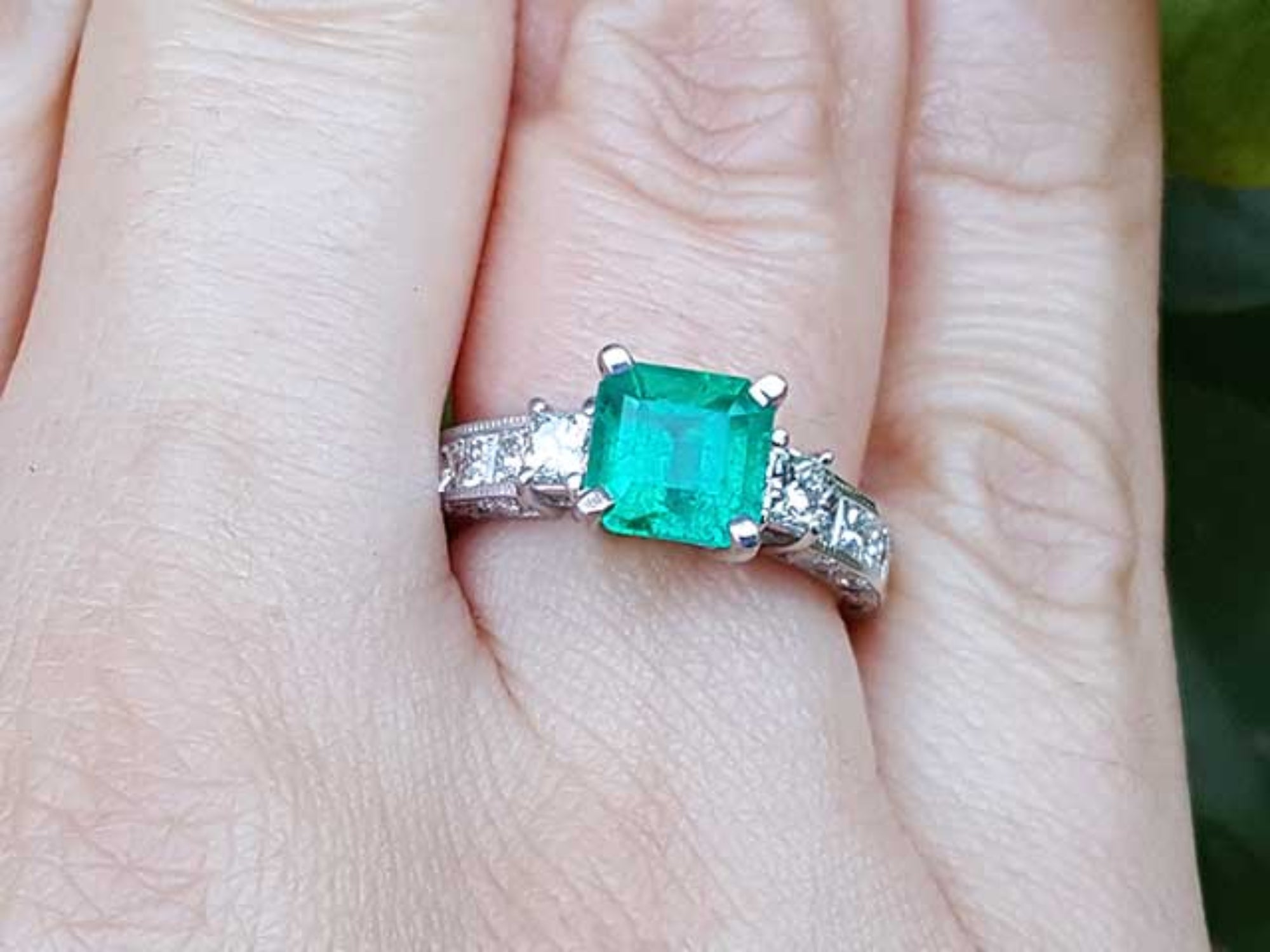 Hand wearing a square emerald ring with side diamond princess cut in white gold.
