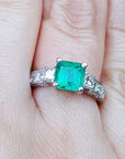 Hand wearing a square emerald ring with side diamond princess cut in white gold.