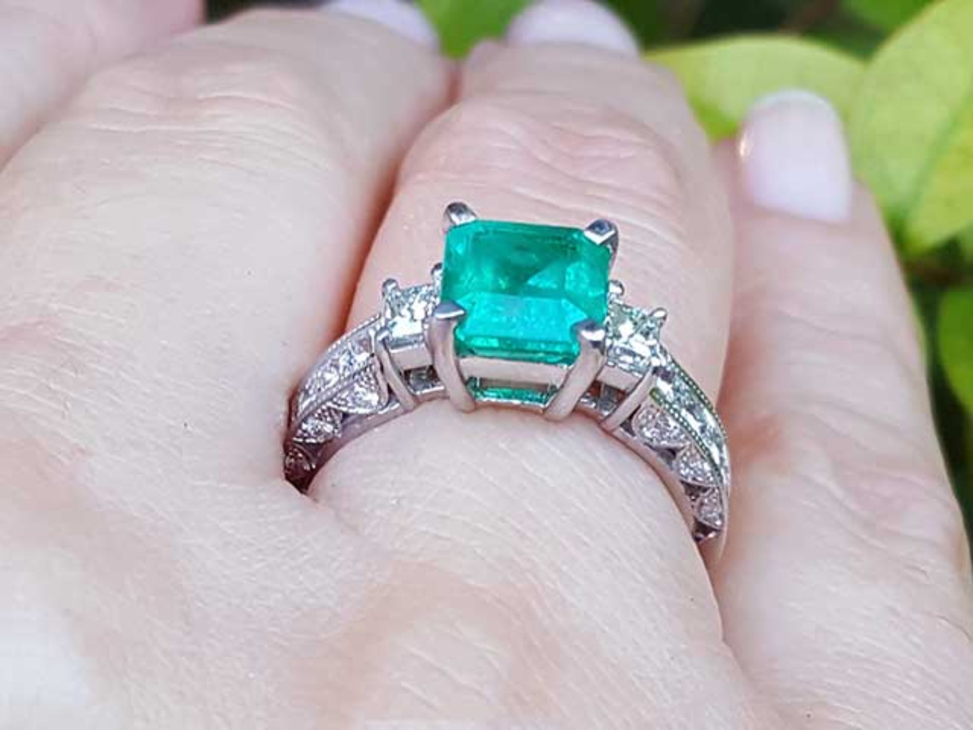 On a woman's hand a square Colombian emerald engagement ring, against a leafy background.
