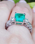 On a woman's hand a square Colombian emerald engagement ring, against a leafy background.