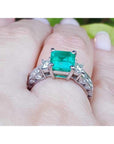 On a woman's hand a square Colombian emerald engagement ring, against a leafy background.
