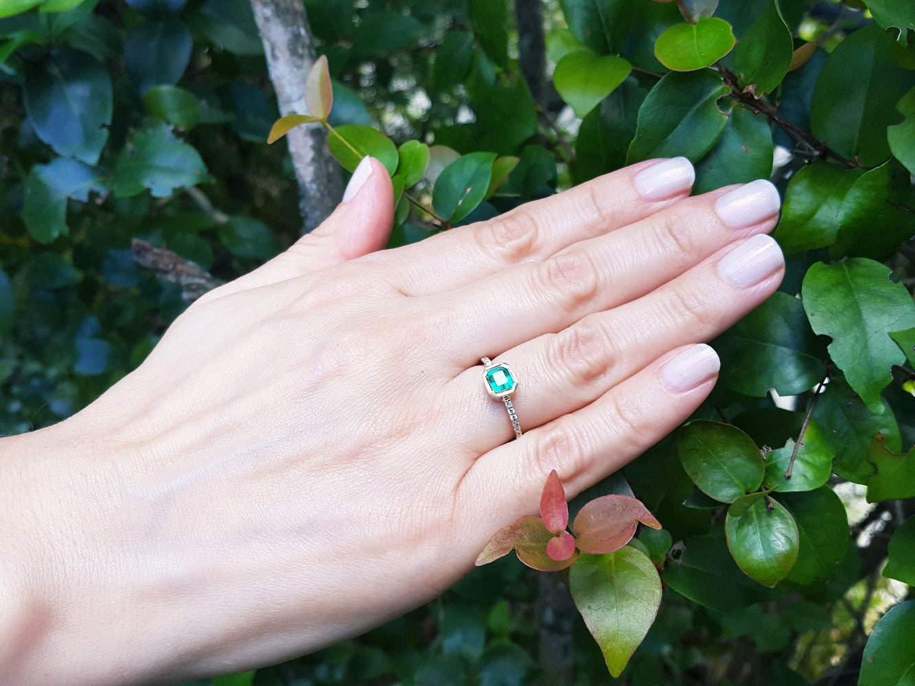 Hand wearing a colombian emerald ring against a leafy green background.