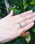 Hand wearing a colombian emerald ring against a leafy green background.