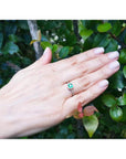 Bezel-set emerald ring with accent diamonds in 14K yellow gold, worn on a hand amidst green leaves.