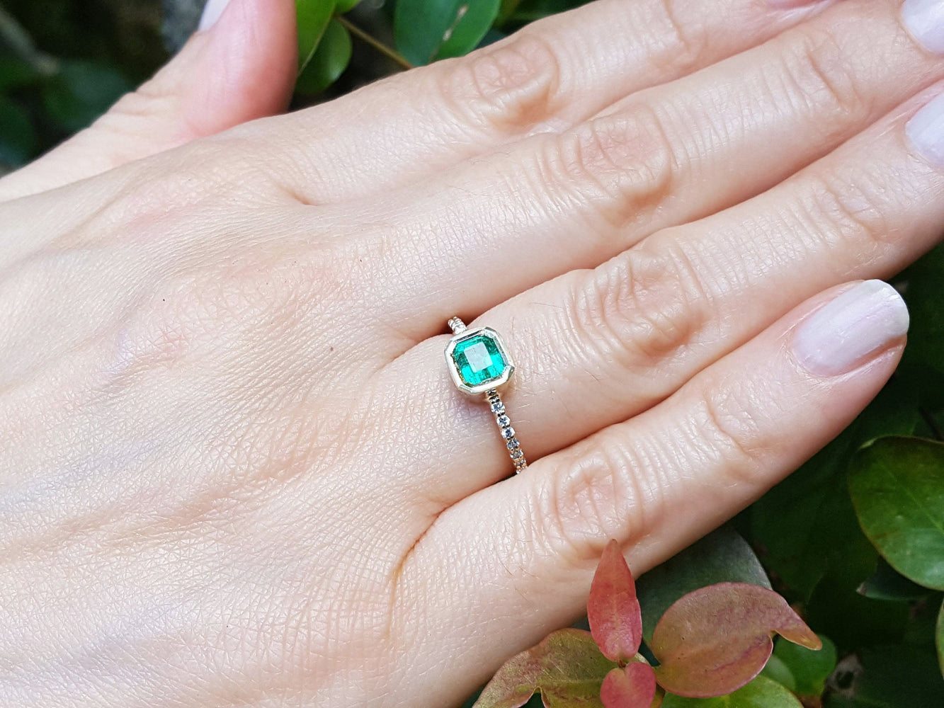 Hand wearing a bezel emerald set ring accent diamonds against a natural background.