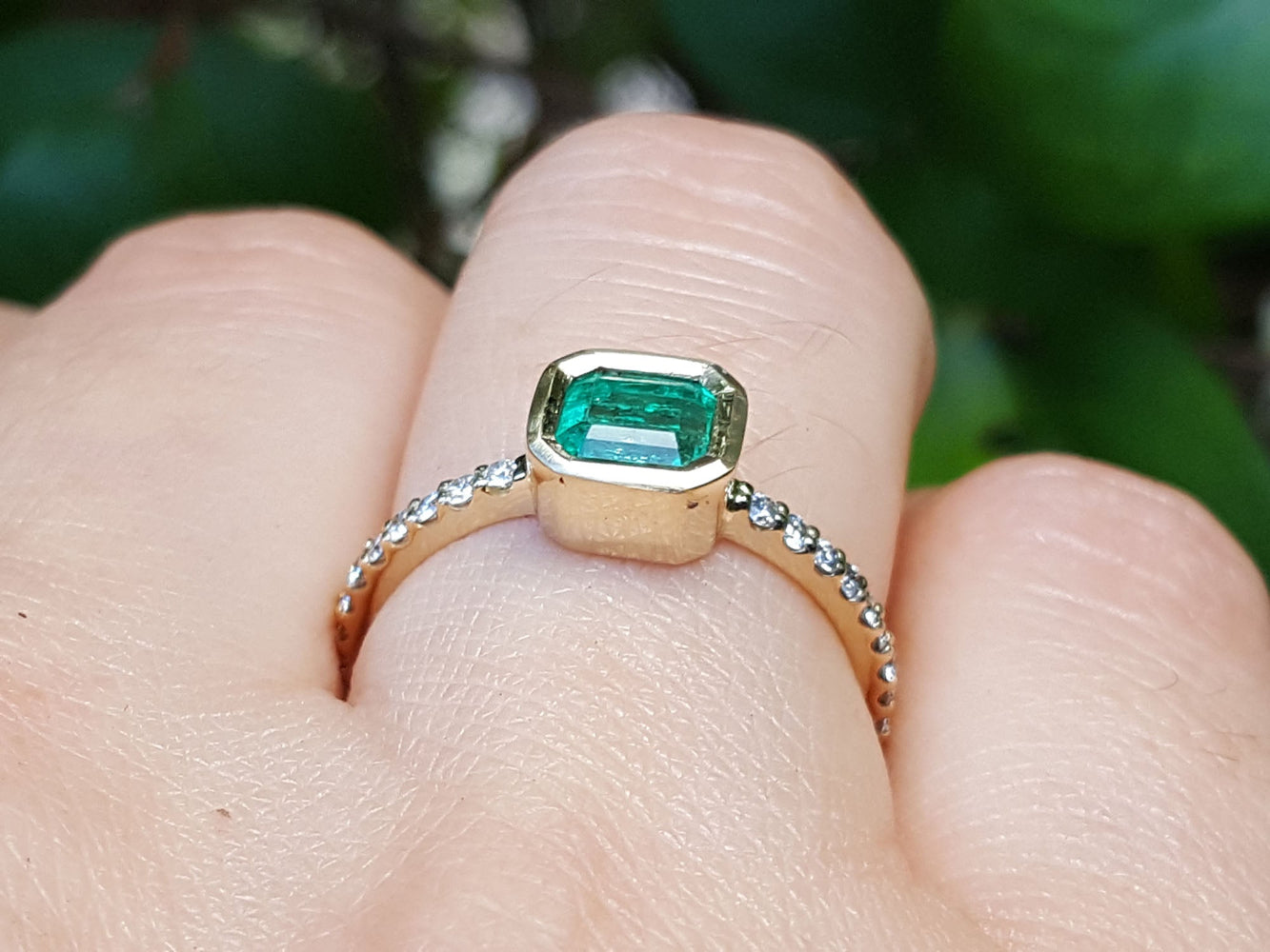 A yellow gold emerald ring on a finger against a blurred natural background.