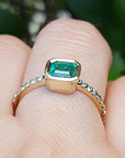 A yellow gold emerald ring on a finger against a blurred natural background.
