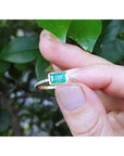 A yellow gold emerald ring being held between fingers against a leafy background.