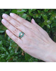 Hand wearing a 14K yellow gold emerald ring guard with a solitaire diamond ring, against green foliage.