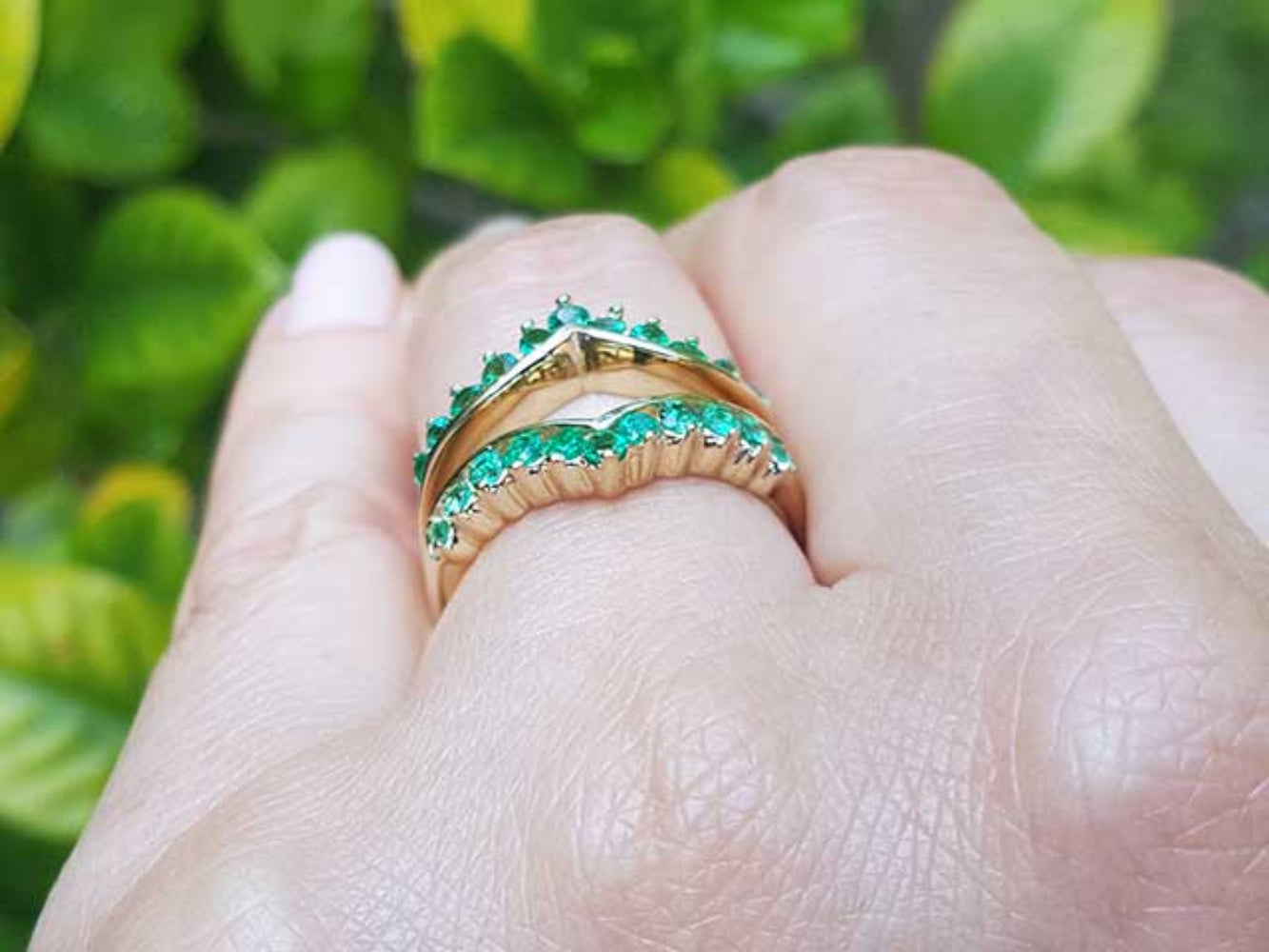 A yellow gold emerald ring guard on a hand against a blurred green leafy background,