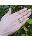 Hand wearing a ring with an emerald stone against a green leafy background.