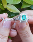 Close-up of a hand holding an emerald ring with matching band against a blurred green leafy background.