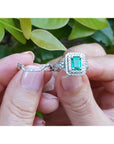 Close-up of a hand holding an emerald ring with matching band against a blurred green leafy background.