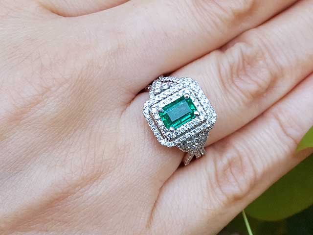 Emerald ring with double halo diamond on a hand against a natural background.