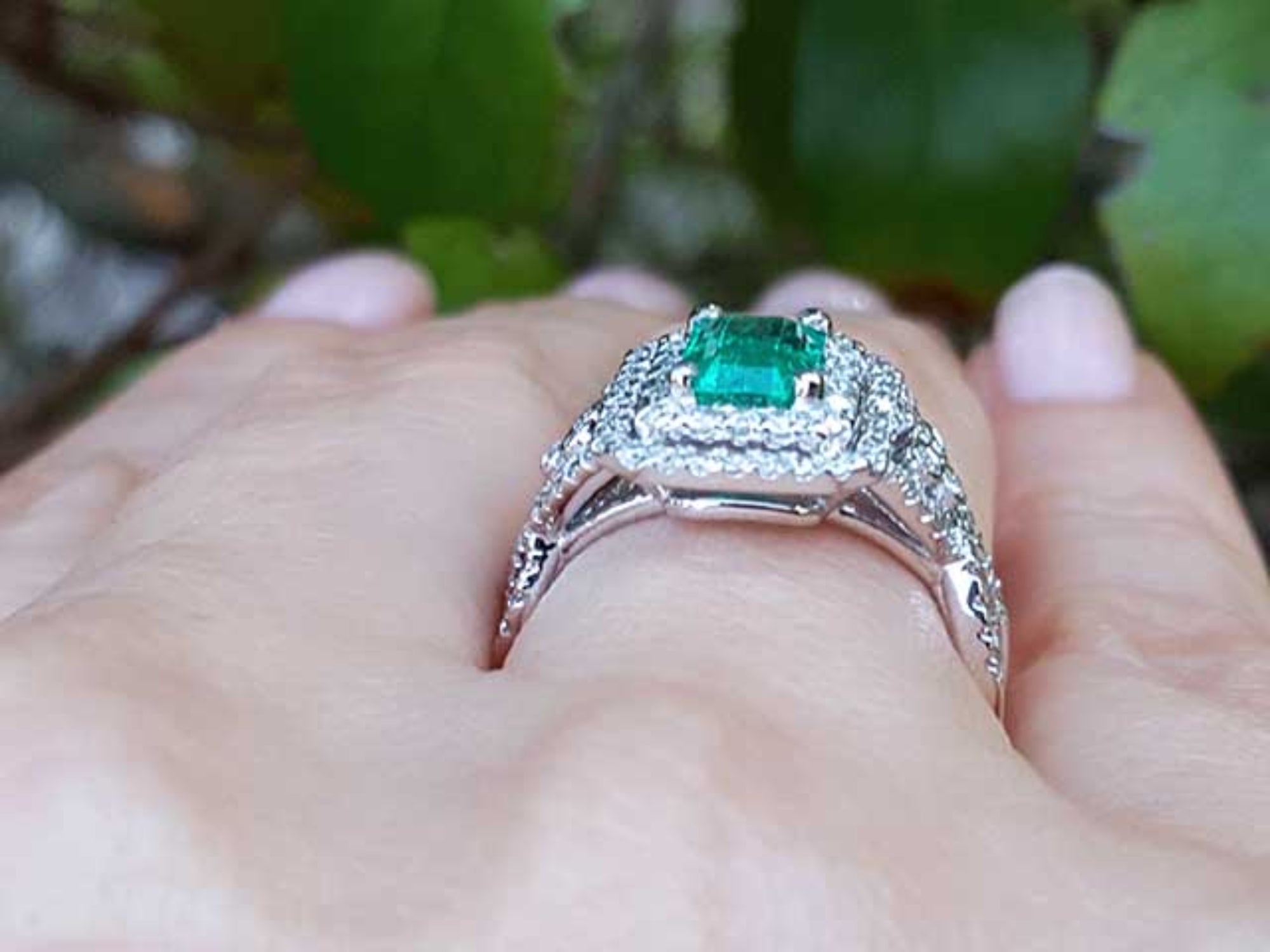 A white gold emerald ring on a hand against a blurred green background.