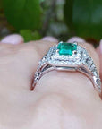 A white gold emerald ring on a hand against a blurred green background.