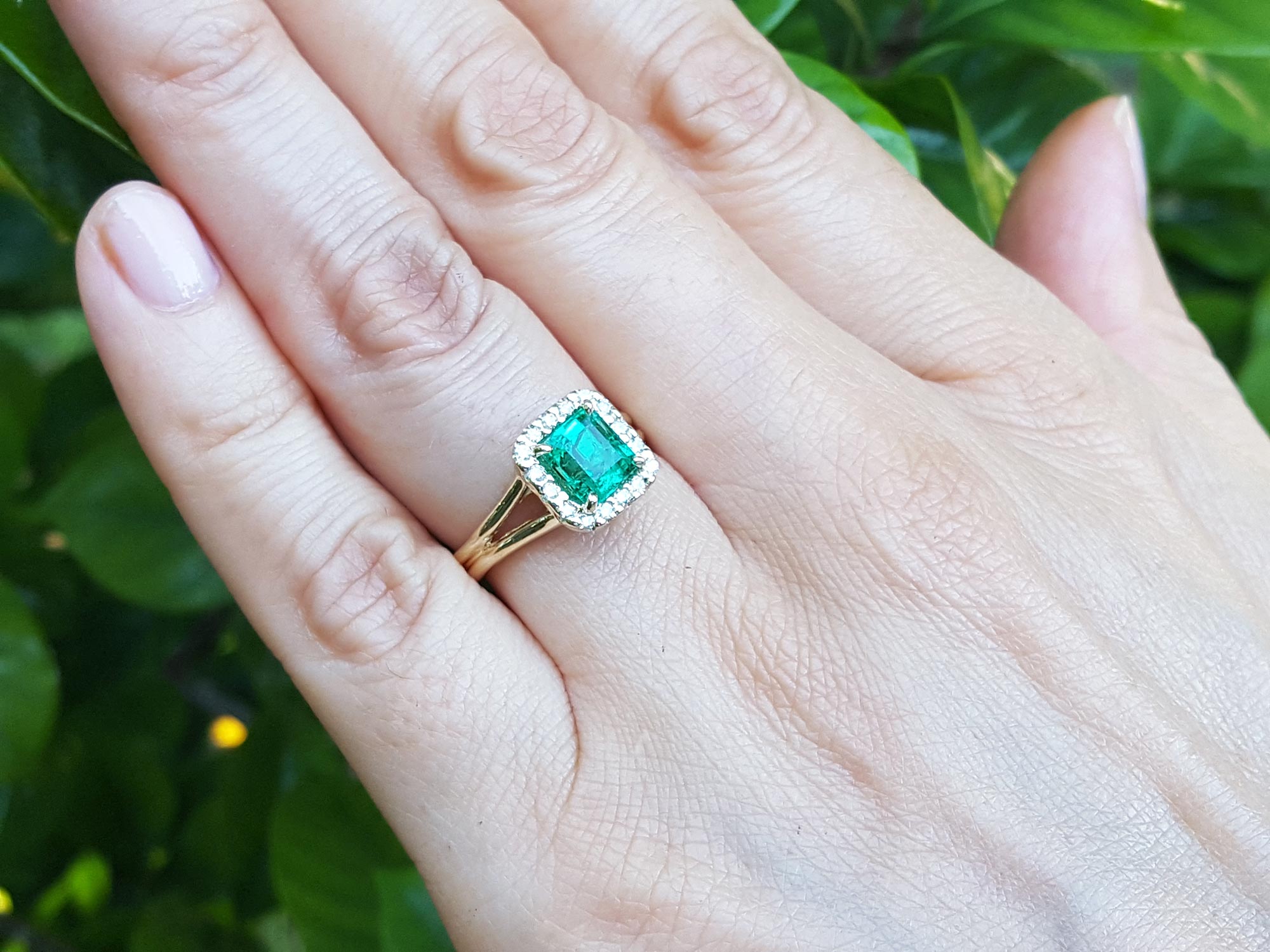 Hand wearing a square emerald ring with halo diamond against a leafy background