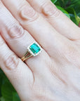 Hand wearing a square emerald ring with halo diamond against a leafy background