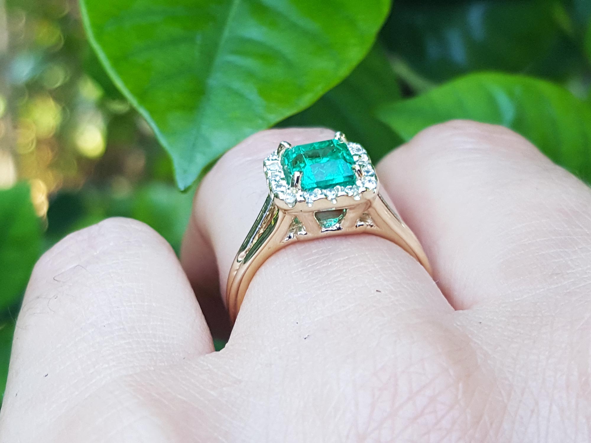 Emerald halo diamond ring underside view on a finger over green leaves background.