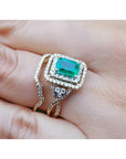 1.31 ct. emerald-cut emerald ring with double diamond halo and matching 14K yellow gold band on a finger.