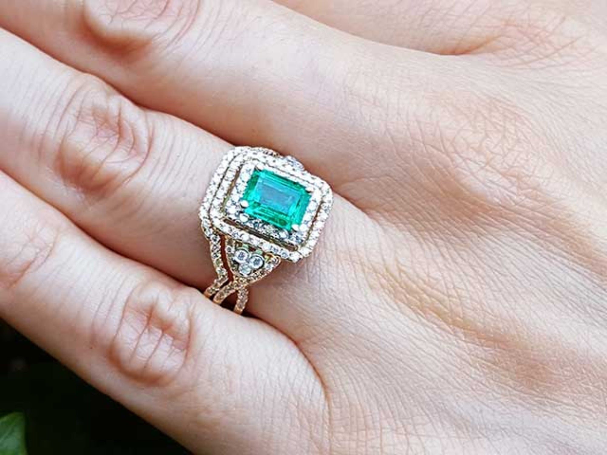 Ring with an emerald-cut emerald within a double halo diamond on a hand.