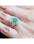 Ring with an emerald-cut emerald within a double halo diamond on a hand.