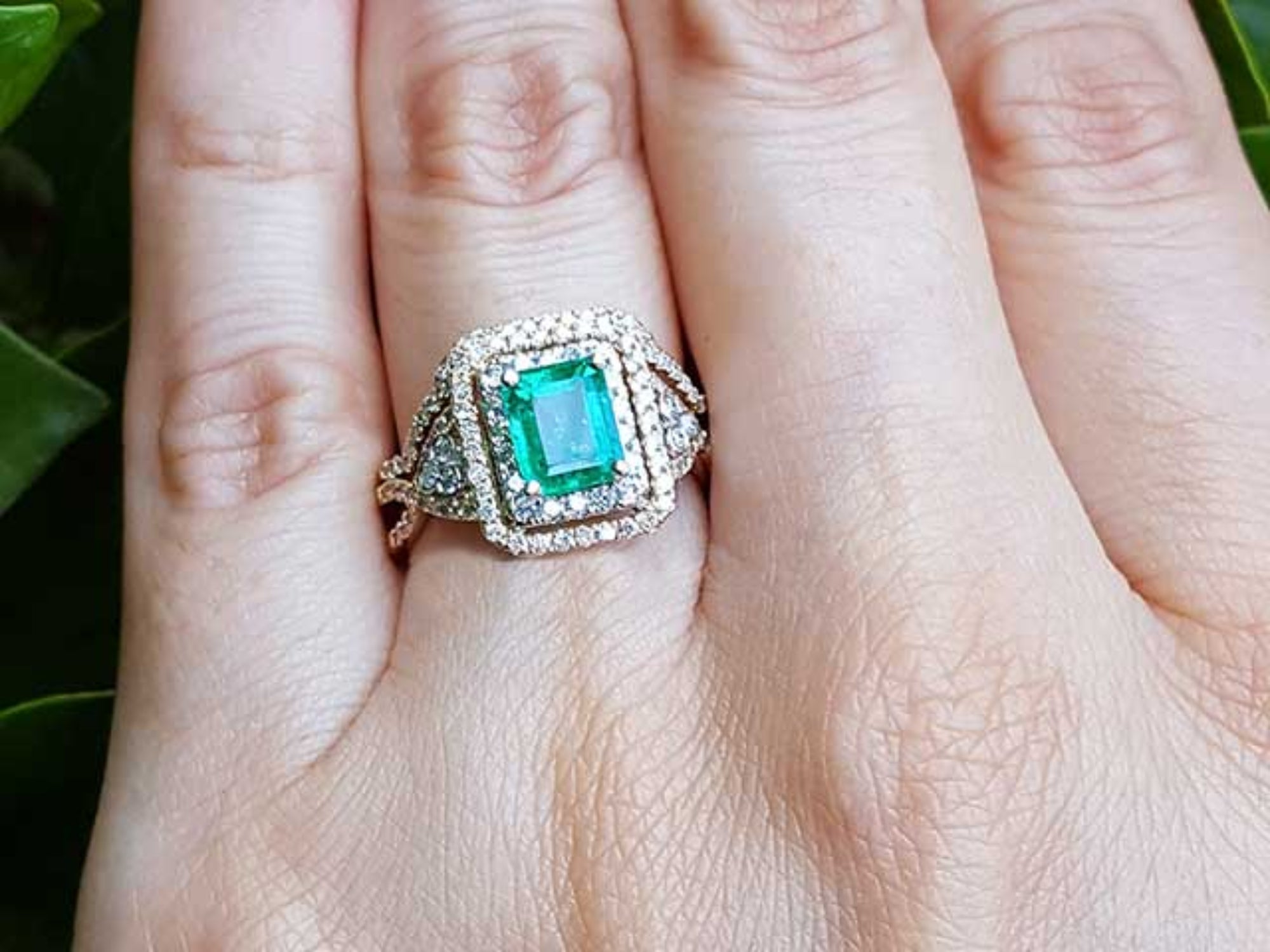 A bridal emerald engagement ring on a hand against a natural background.