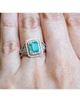 A bridal emerald engagement ring on a hand against a natural background.