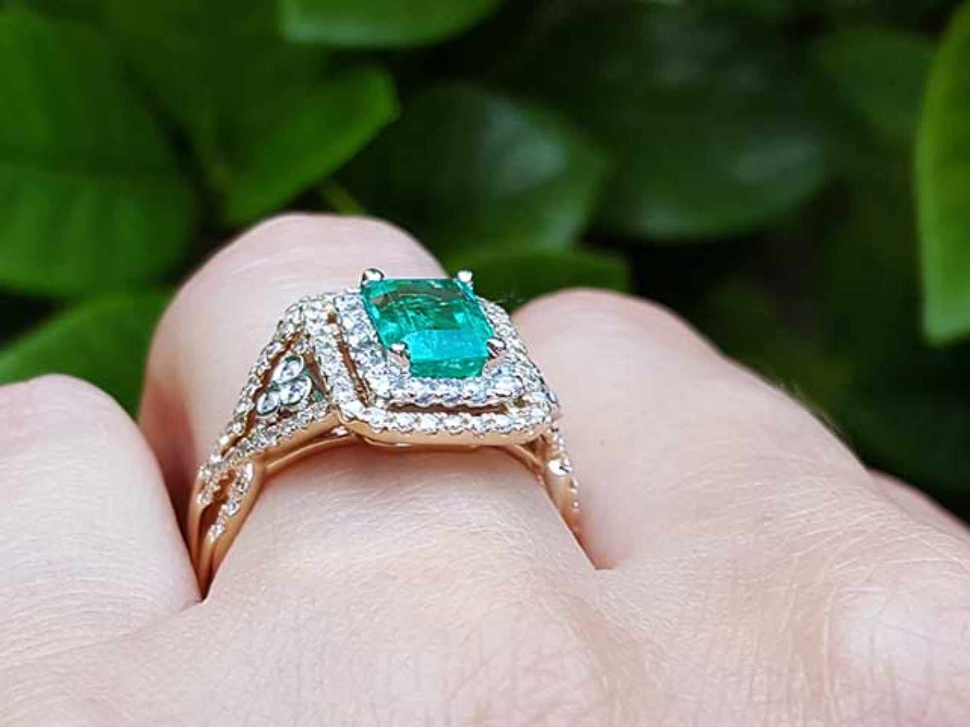 Yellow gold ring with emerald and diamonds on a hand against a green leafy background.