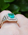Yellow gold ring with emerald and diamonds on a hand against a green leafy background.