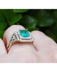 Yellow gold ring with emerald and diamonds on a hand against a green leafy background.