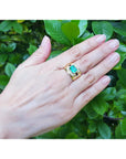 Hand wearing a gold men's emerald ring bezel set against a green leafy background.