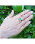 Hand wearing a gold men's emerald ring bezel set against a green leafy background.