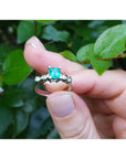 Hand holding a natural emerald ring against a green leafy background.