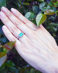 Hand wearing a natural emerald ring with accent diamonds against a green leafy background.