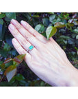 Emerald-cut emerald ring with natural diamonds on a 14K white gold band, worn on a hand amidst green leaves.