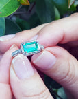 Close-up of a natural emerald ring in white gold held between fingers against a blurred green leaf background.