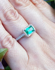 Emerald ring on a hand with green leaves in the background.