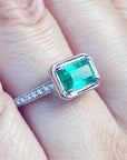 A white gold emerald ring with accent natural diamonds on a finger.